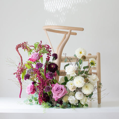 A floral arrangement with a split palette sits on the floor in front of a wooden chair. The chair is in profile, and the flowers are purple and white.